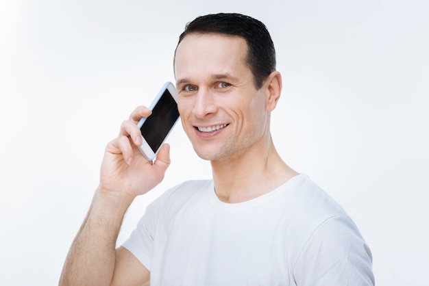 Llamada telefónica. Hombre agradable agradable alegre sosteniendo su teléfono inteligente y mirándote mientras haces una llamada telefónica