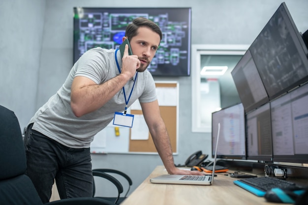 Foto llamada urgente. hombre concentrado mirando de cerca los monitores y hablando por teléfono inteligente inclinado sobre la mesa