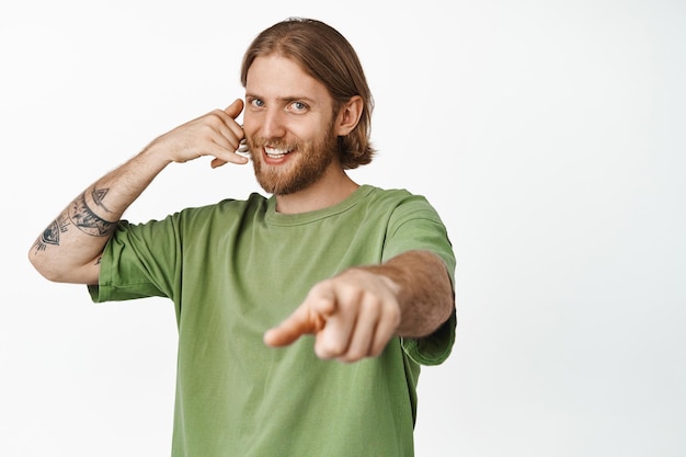 Llámame. Hombre barbudo sonriente que muestra un gesto de teléfono móvil cerca de la oreja, apuntándote, coqueteando, diciéndole que lo contactes, parado sobre fondo blanco