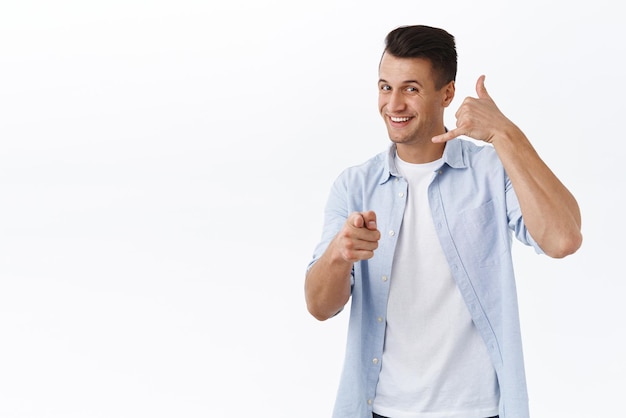 Llámame Retrato de un apuesto y alegre chico caucásico que muestra la señal del teléfono y apunta con la cámara quiere tener una conversación hablar con la persona que coquetea contigo de fondo blanco