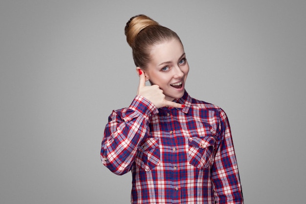 ¡Llámame! retrato de una chica rubia hermosa y graciosa con camisa rosa a cuadros, peinado de moño recogido y maquillaje de pie y mirando a la cámara con gesto de llamada. interior, aislado sobre fondo gris