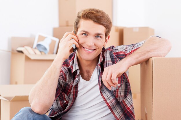 Llamando desde casa nueva. Apuesto joven sentado en el suelo y hablando por teléfono móvil mientras cajas de cartón en el fondo