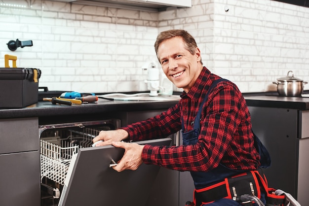 Llame al técnico masculino profesional sentado cerca del lavavajillas y sonriendo