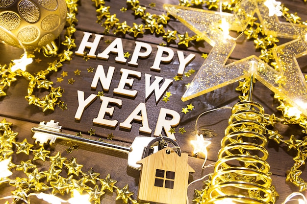 Llave de casa con llavero cabaña sobre un fondo festivo con lentejuelas, estrellas, luces de guirnaldas. Feliz año nuevo-letras de madera, saludos, tarjetas de felicitación. Compra, construcción, reubicación, hipoteca
