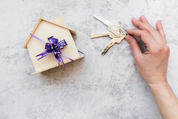 Las llaves de una mano de una persona cerca del modelo de casa de madera con un lazo morado sobre un fondo de concreto