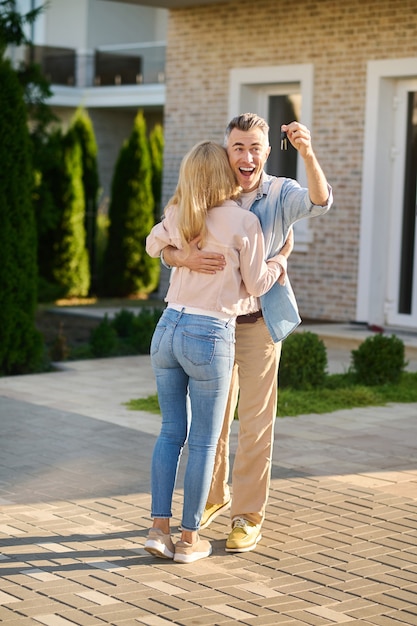 Llaves, nueva casa. Hombre adulto joven entusiasta con la boca abierta mostrando llaves abrazando a esposa de pelo largo cerca de la nueva casa en la buena tarde