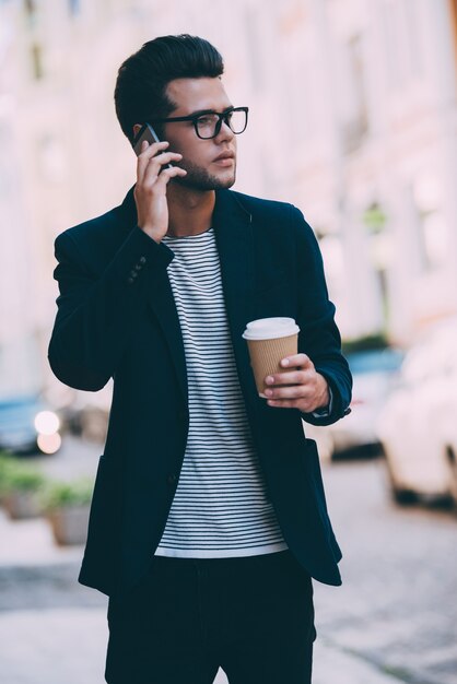 Llegaré a tiempo. Apuesto joven en ropa casual elegante con taza de café y hablando por teléfono móvil mientras camina por la calle