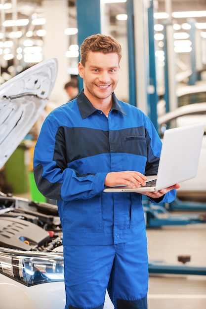 Lleno de confianza y experiencia. Hombre joven confidente que trabaja en la computadora portátil y sonriendo mientras está de pie en el taller con el coche en el fondo