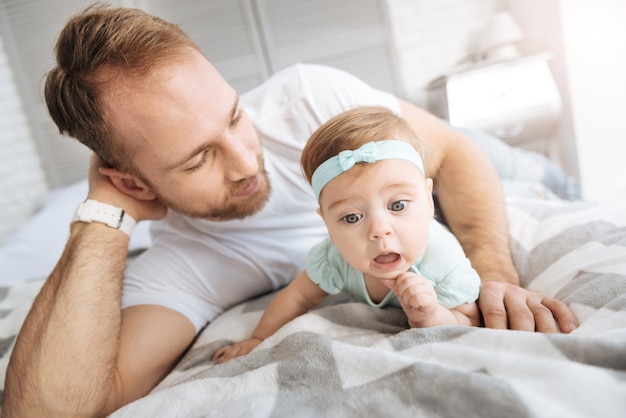 Lleno de diferentes emociones. Encantadora asombrada encantadora niña pequeña acostada en la cama con su padre y relajándose mientras expresa interés