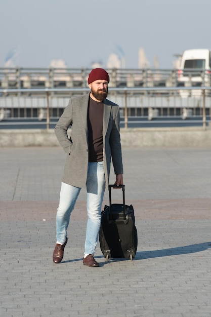 Lleva bolsa de viaje. Hombre barbudo hipster viaja con bolsa de equipaje sobre ruedas. El viajero con maleta llega a la estación de tren del aeropuerto de fondo urbano. Mudarse a la nueva ciudad solo. Hipster listo para disfrutar de los viajes.