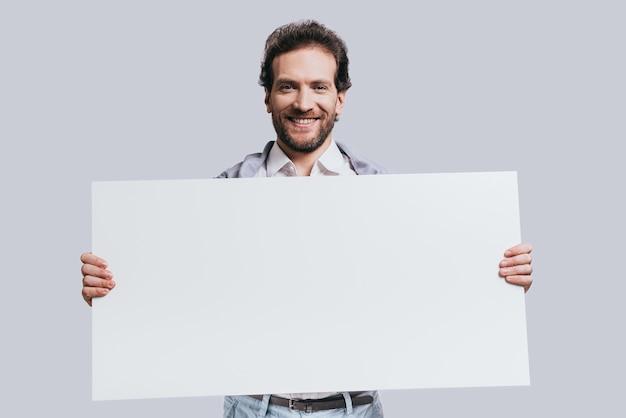 Llevando copia espacio. Hombre confiado joven en ropa casual elegante que sostiene el rotafolio en blanco y sonriendo mientras está de pie