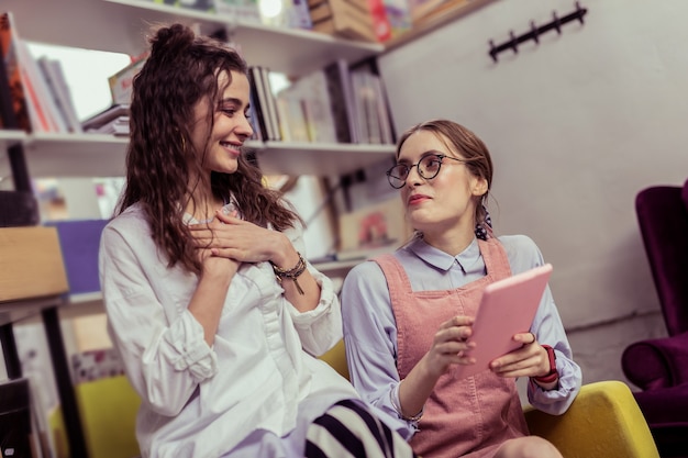 Llevando portátil rosa. Atractivas damas positivas que tienen una conversación agradable y agradable mientras están sentadas en un área para relajarse