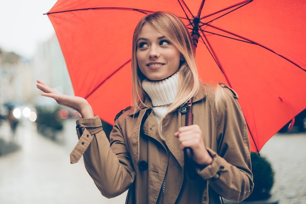 ¿Esta lloviendo? Atractiva mujer joven con paraguas y estirando la mano mientras está de pie en la calle