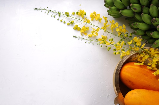 Lluvia dorada pepino flor amarilla y racimo de plátano crudo dispuestos sobre una superficie blanca con fondo de textura gris y un recipiente tradicional de latón o urule dispuestos en el fondo