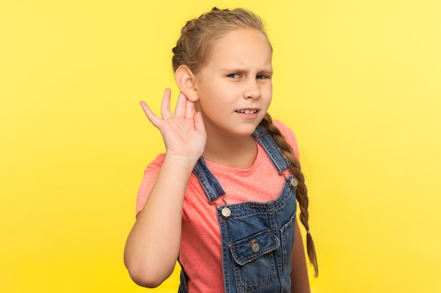 Lo que no puedo escuchar Retrato de una niña curiosa y atenta con overoles de mezclilla sosteniendo la mano cerca de la oreja y escuchando atentamente lo que dices en un estudio interior aislado en un fondo amarillo