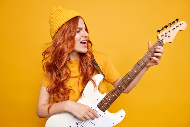 Loca y alegre artista femenina realiza música rock en una guitarra acústica eléctrica, usa un sombrero y una camiseta tiene el pelo rojo natural y canta una famosa canción legendaria que posa contra una pared amarilla vibrante. Entretenimiento