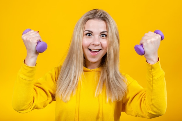 Loca hermosa rubia joven en ropa deportiva amarilla casual haciendo ejercicios con pequeñas pesas moradas de 0.5 kg.