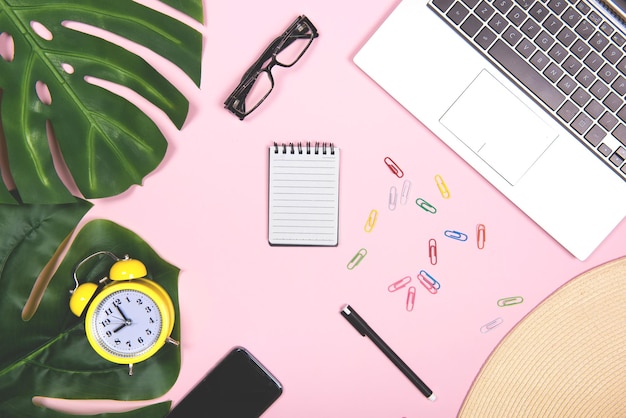 Local de trabalho de escritório com caneta de telefone planejador diário de computador portátil e óculos com decoração tropical feriados de negócios plano para mídias sociais em fundo rosa