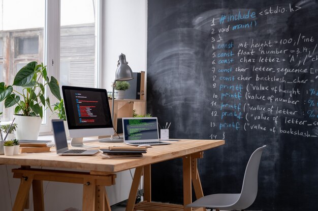 Local de trabalho do engenheiro de ti pela janela do escritório com dois laptops, monitor de computador, alguns livros e cadernos, abajur e plantas domésticas