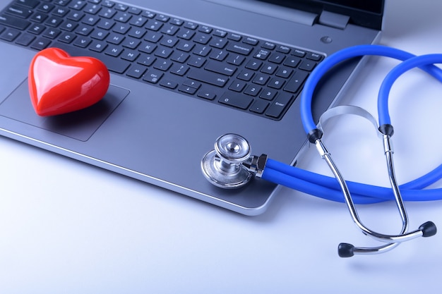 Foto local de trabalho do médico com laptop, estetoscópio, coração vermelho e caderno na mesa branca.