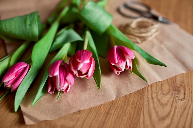 Local de trabalho florístico com papel ofício, barbante, Arranjando buquê de tulipas cor de rosa na mesa de madeira, hobby, faça você mesmo, conceito de presente de primavera, de cima.