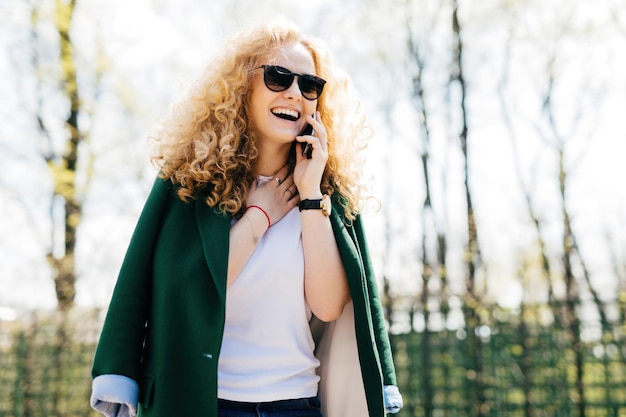 Lockenhaarige Frau in stilvoller Kleidung und Sonnenbrille, die auf dem Smartphone spricht, lächelt und lacht vor Aufregung, glücklicher Gesichtsausdruck, Nackenberührung
