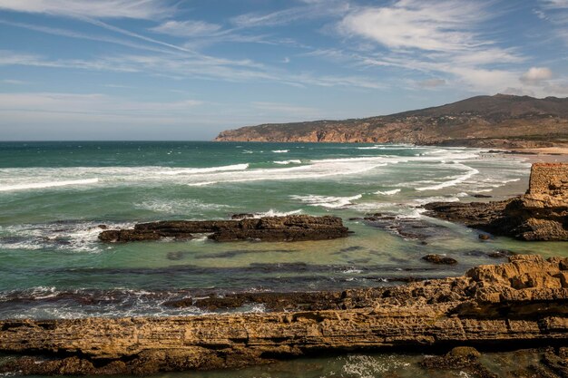 Lockere Felsklippen und anhaltende Wellen an der portugiesischen Atlantikküste und ein Strand mit Sonnenanbetern an einem großartigen Sommertag