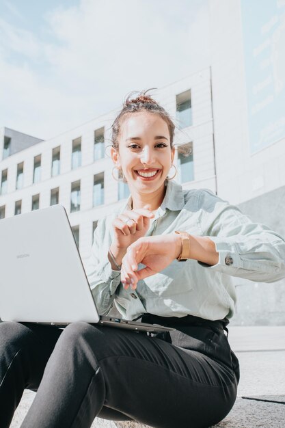 Lockige afrikanische Manager-Geschäftsfrau, die lächelnd auf die Kamera schaut, mit Laptop, der auf der Treppe sitzt. Tragende weiße Bluse der attraktiven jungen Geschäftsfrau. Gruß Textfreiraum