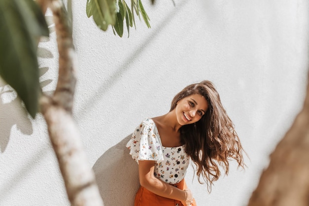 Lockige Frau in toller Stimmung posiert auf weißem Hintergrund mit Bäumen Lady in white floral print top lächelnd