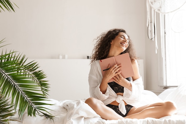 lockige junge Frau in Dessous, die lächelt und ein Tagebuch auf dem Bett in einer weißen Wohnung hält