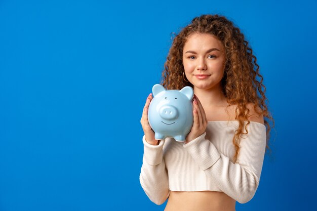 Lockige junge Frau mit Sparschwein vor blauem Hintergrund