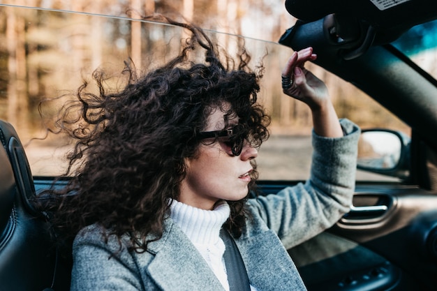 Lockige schöne Frau in einem Cabrio an einem sonnigen Tag mit Sonnenbrille entspannt sich und fühlt sich frei