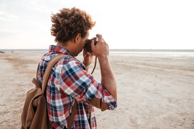 Lockiger afroamerikanischer junger Mann mit Rucksack, der am Strand steht und fotografiert