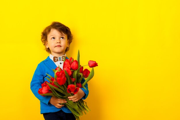 lockiger lustiger kleiner Junge in Jacke und Fliege mit einem Strauß Tulpen in den Händen schaut auf