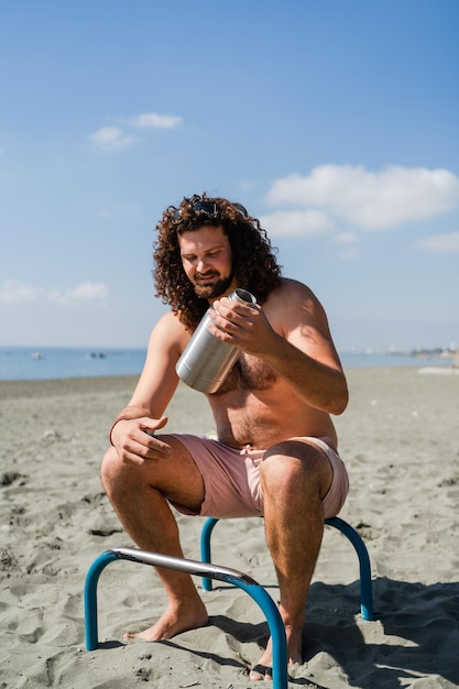 Lockiger Mann entspannt sich und trinkt Wasser nach dem Training am Strand