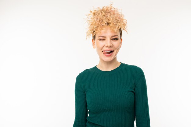 lockiges blondes Mädchen in grüner Kleidung zeigt Zunge auf Weiß mit Copyspace