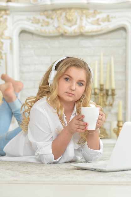 Lockiges blondes Teenager-Mädchen mit Kopfhörern, das mit einer Tasse Tee und offenem Laptop auf dem Boden liegt