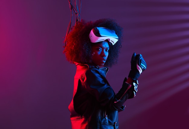 Lockiges dunkelhaariges Mädchen in schwarzer Lederjacke und Handschuhen trägt die Virtual-Reality-Brille auf dem Kopf im dunklen Studio mit Neonlicht. Cyberpunk-Charakter