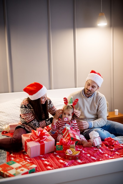 Lockiges kleines Mädchen mit weihnachtlichen Rentierohren und Schnuller, die mit ihren vorsichtigen Eltern und Geschenken auf dem Bett sitzen.