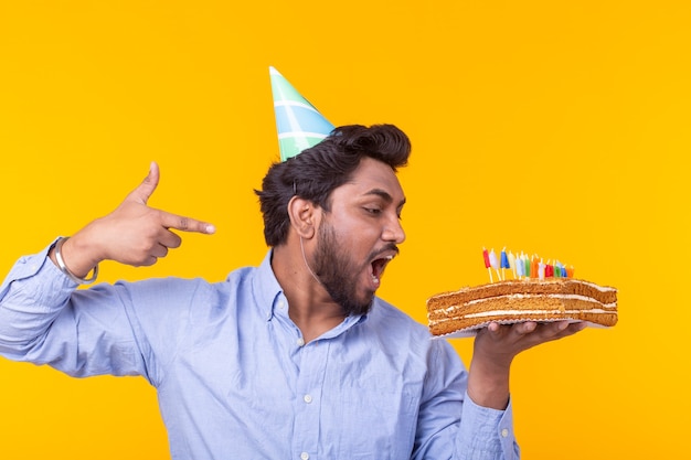 Foto loco alegre joven indio con sombrero de papel de felicitación sosteniendo pasteles feliz cumpleaños de pie sobre una superficie amarilla. concepto de felicitaciones de jubileo.