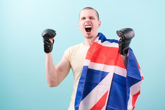 Un loco luchador de MMA gritando con guantes negros de pie sobre un fondo azul y sostiene una bandera del Reino Unido Pesado Duro Bueno Satisfecho Golpear Ganar Gol Condición Amenaza Alegre Positivo