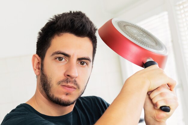 Loco muy enojado joven amenazando con sartén en la cocina.