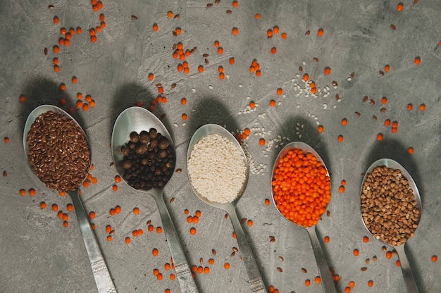 Löffel mit verschiedenen Superfoods - Leinsamen, Sesam, Pfeffer, rote Linsen, Buchweizen auf einer grauen Oberfläche. Gesundes veganes Essen.