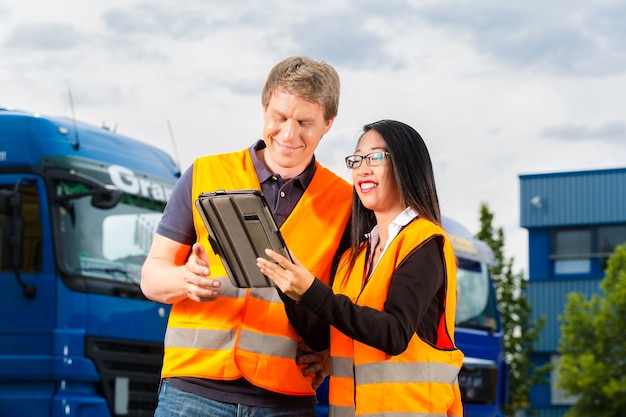 Logística - motorista orgulhosa ou despachante e colega de trabalho com tablet, na frente de caminhões e reboques, em um ponto de transbordo, é uma equipe boa e bem-sucedida