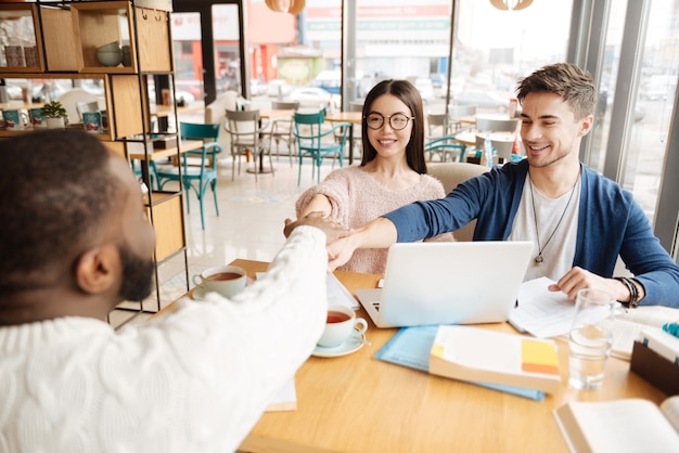 Lograremos metas. Tres jóvenes estudiantes internacionales atractivos se dan la mano mientras están sentados en la cafetería.