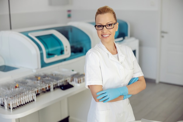 Loira atraente sorridente assistente de laboratório em pé no laboratório com as mãos cruzadas.