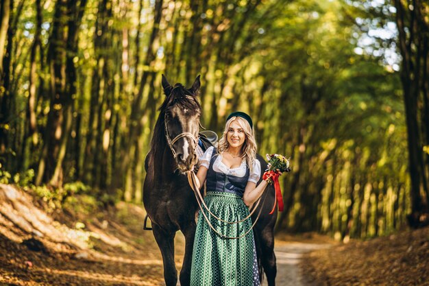 Loira bonita no vestido tradicional andando com grande cavalo preto na floresta