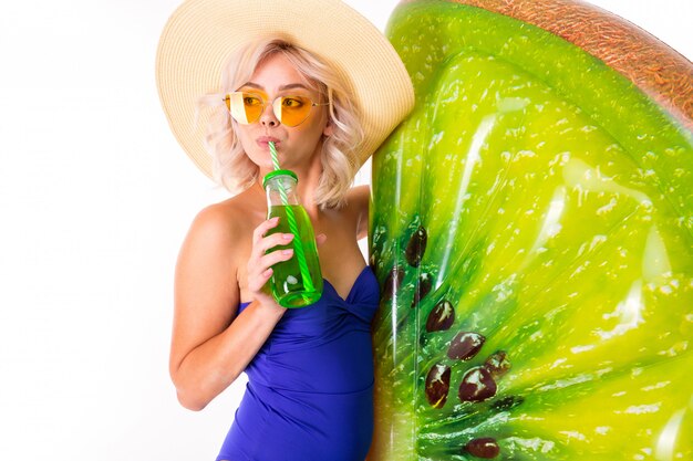 Loira caucasiana bonita fica em traje de banho com colchão de kiwi de praia de borracha, beber suco e sorrisos isolados no fundo branco