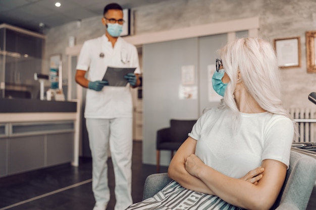 Loira com máscara facial sentada no corredor com os braços cruzados e esperando os resultados dos testes para coronavírus.