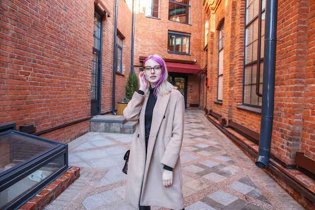 Foto loira com mechas de cabelo lilás em um casaco bege e óculos no fundo de uma construção de tijolo vermelho